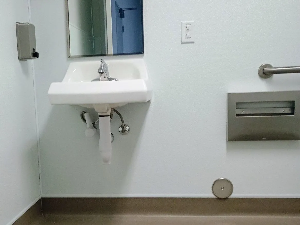 Wall-mount sink with a single-lever faucet under a mirror in a white-paneled restroom, a grab bar and a paper towel dispenser on the wall to the right