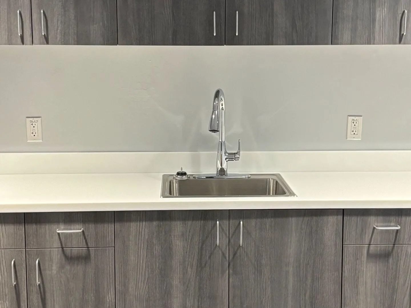 Stainless break room sink with a pull-down faucet set in a white counter between grey wood cabinets