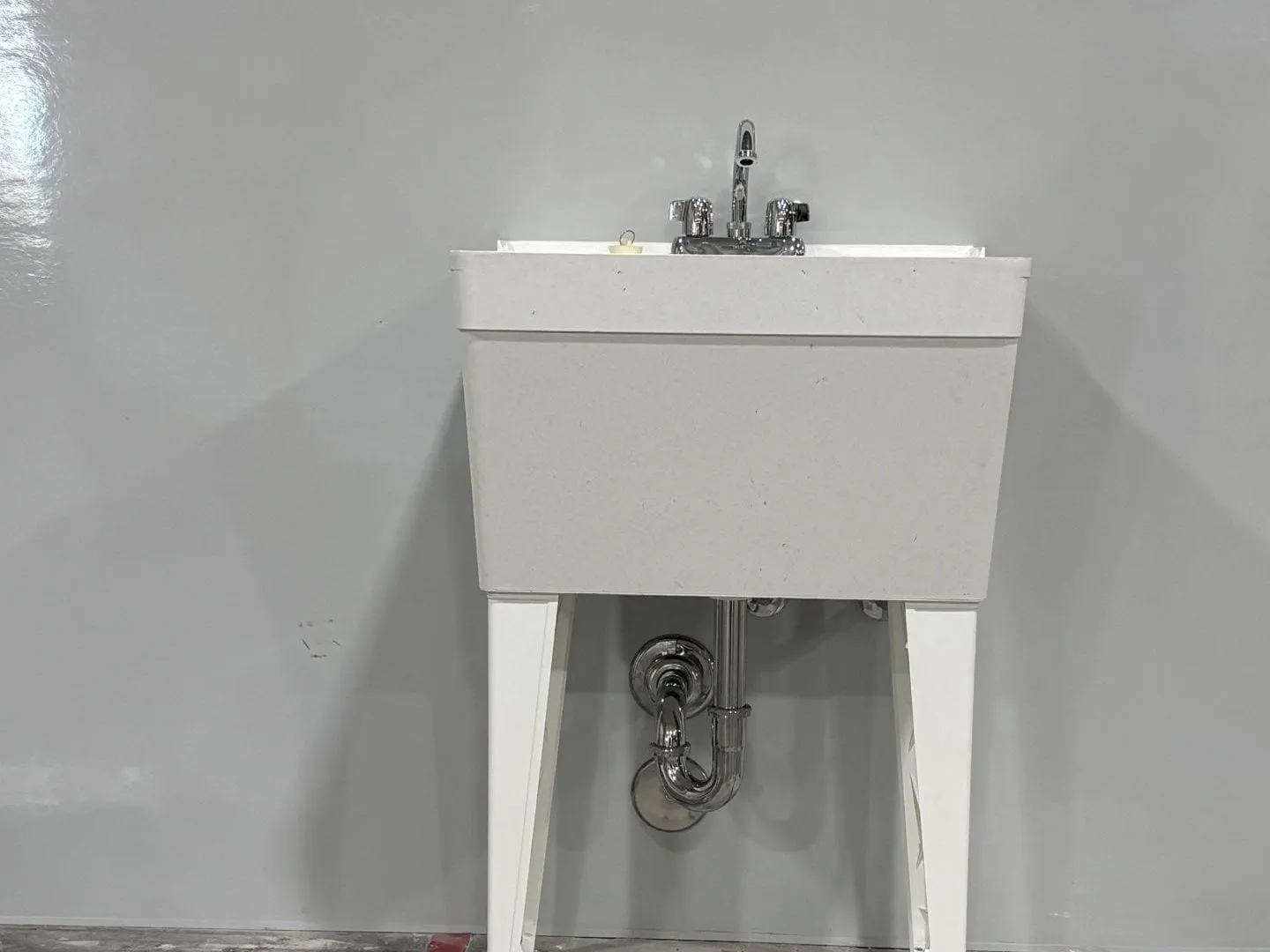 A utility-tub janitor's sink on white legs against a grey warehouse wall, with a two-handle faucet and a chrome trap