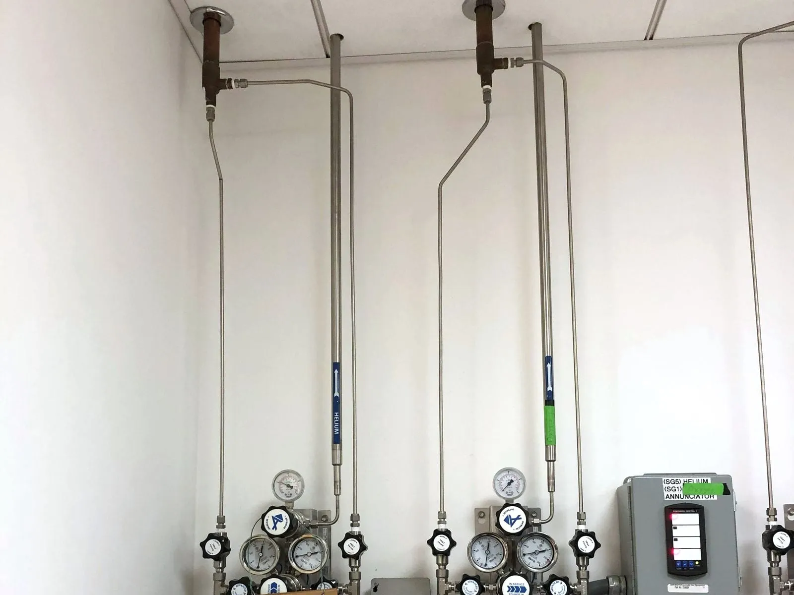 Labeled stainless helium lines running down a white wall from the ceiling to the cylinder regulators
