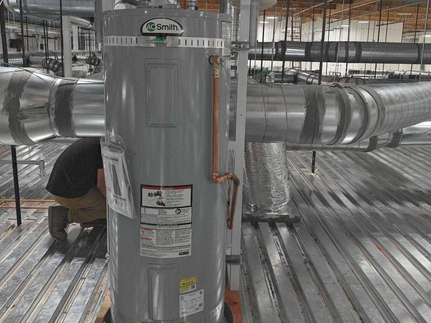 Electric water heater standing in its drain pan on a mezzanine deck among ducts, a worker kneeling behind it