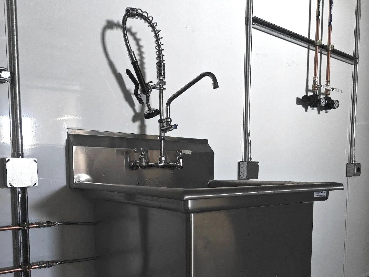 Stainless sink with a pre-rinse faucet and a second spout, copper lines on the wall beside it
