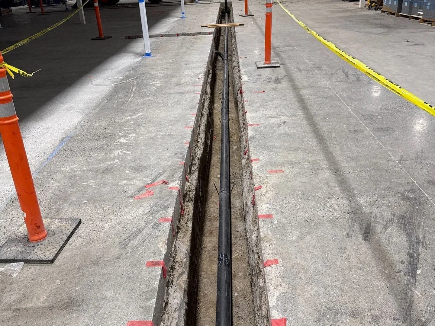 A long trench cut in a warehouse floor with black waste pipe laid in it, cones and caution tape alongside