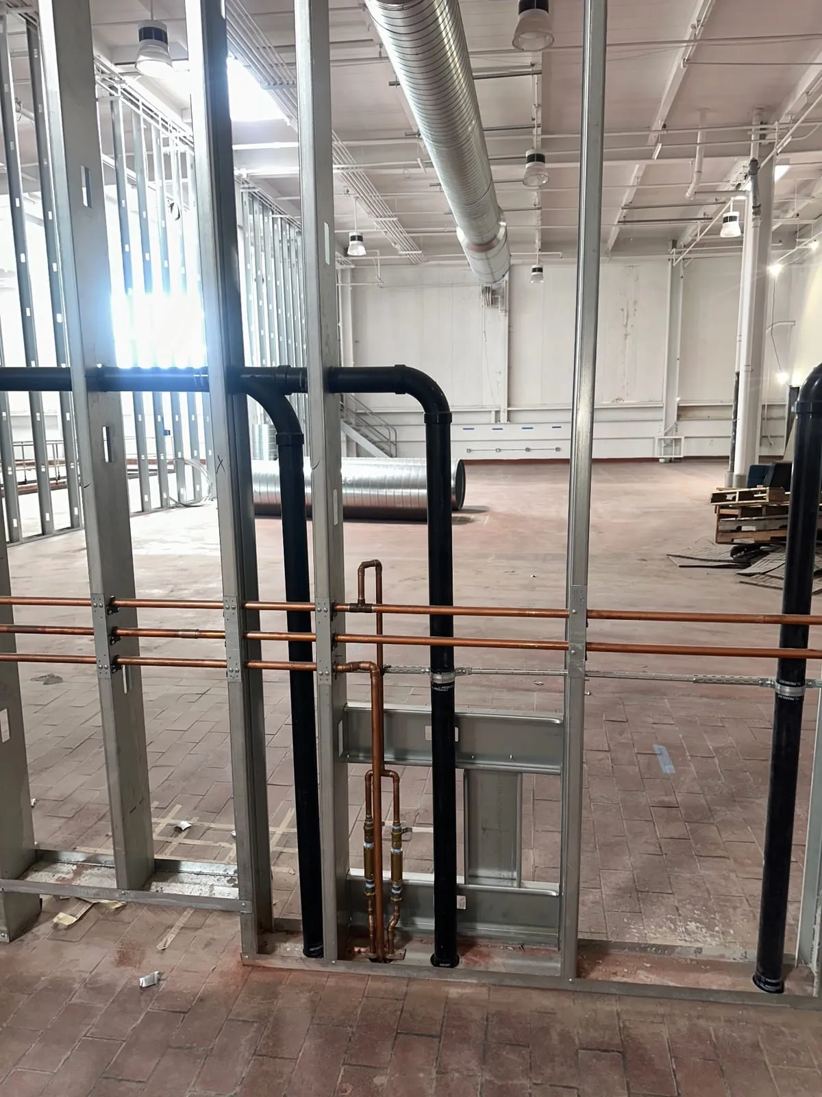 Copper water lines and black waste piping through open steel framing, warehouse floor beyond